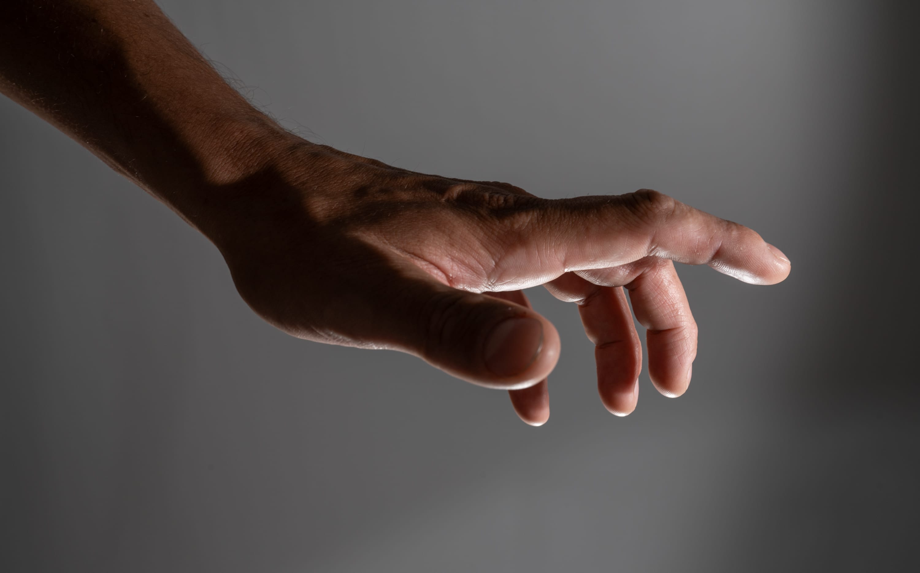 a closeup side view of a man's outstretched hand and arm against a neutral gray background. the hand is reaching forward, fingers slightly curled. the studio lighting creates a dramatic effect, highlighting the contours of the hand and arm.