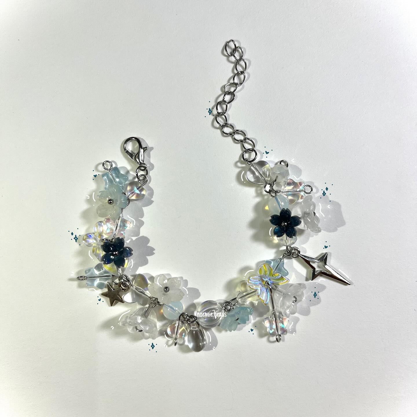 a closeup shot of a handmade bracelet on a white surface. the bracelet features clear and translucent white beads, blue flower beads, silver star charms, and a silver chain. it has a clasp for closure and an additional length of chain for adjusting the size. the overall design is delicate and ethereal.