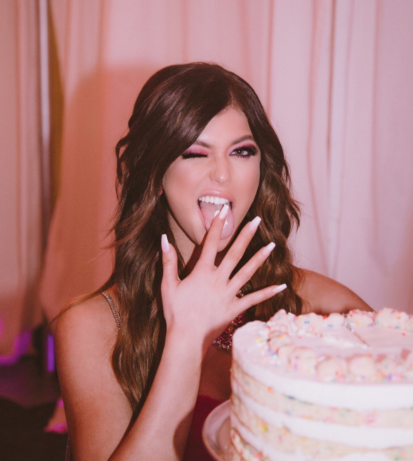 a woman is winking and licking her fingers in front of a birthday cake with sprinkles. she has long nails with white nail polish, brown hair, and pink eyeshadow makeup. she is wearing a pink top. the background is pink.