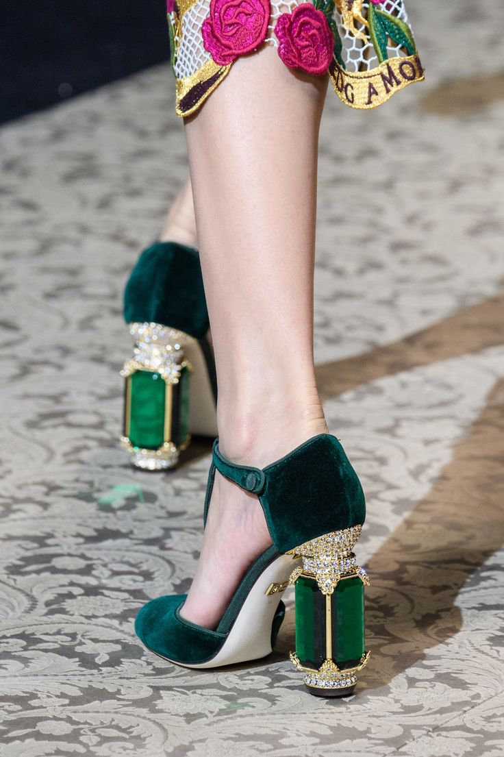 a closeup shot of a model's legs showcasing a pair of exquisite dolce & gabbana high heels. the shoes are made of rich green velvet and feature striking, jewelencrusted block heels resembling emeralds set in gold.  the model is wearing a skirt with floral embroidery visible at the hem. the background is a patterned carpet, typical of a fashion show setting. the image focuses on the intricate detail of the shoes, highlighting their luxurious and glamorous design.