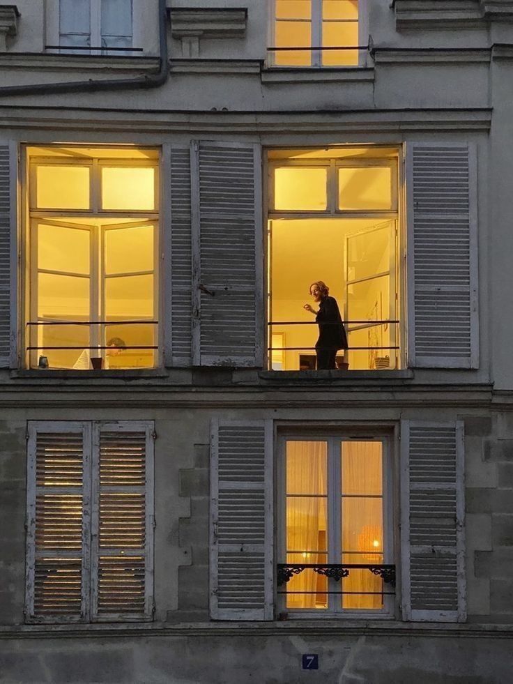 a photograph of a parisian building at night. the building is made of stone, and the windows are lit from within, creating a warm and cozy atmosphere.  the windows are accented with white shutters, and one window shows a woman standing inside, silhouetted against the warm light. the overall aesthetic is vintage and elegant.