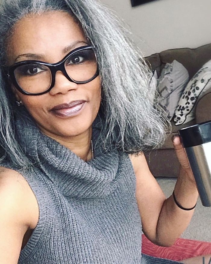 a woman with grey hair is wearing a grey sweater and black glasses. she is holding a silver cup of coffee in her hand.