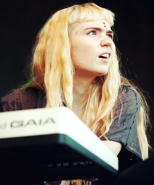 a blonde woman musician with red dots on her forehead is looking up as she is playing a keyboard. she is in a studio and looks focused.  she is wearing a dark gray shirt.  the image is taken close up.
