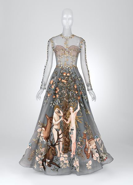 a longsleeved, floorlength gown is displayed on a mannequin against a plain gray backdrop. the gown has a nudecolored underlay at the bodice and sheer fabric covering the rest of the dress. it is intricately embroidered with various designs including trees, branches, flowers, deers, and a scene reminiscent of adam and eve. the embroidery is dense and colorful, creating a detailed and opulent look.