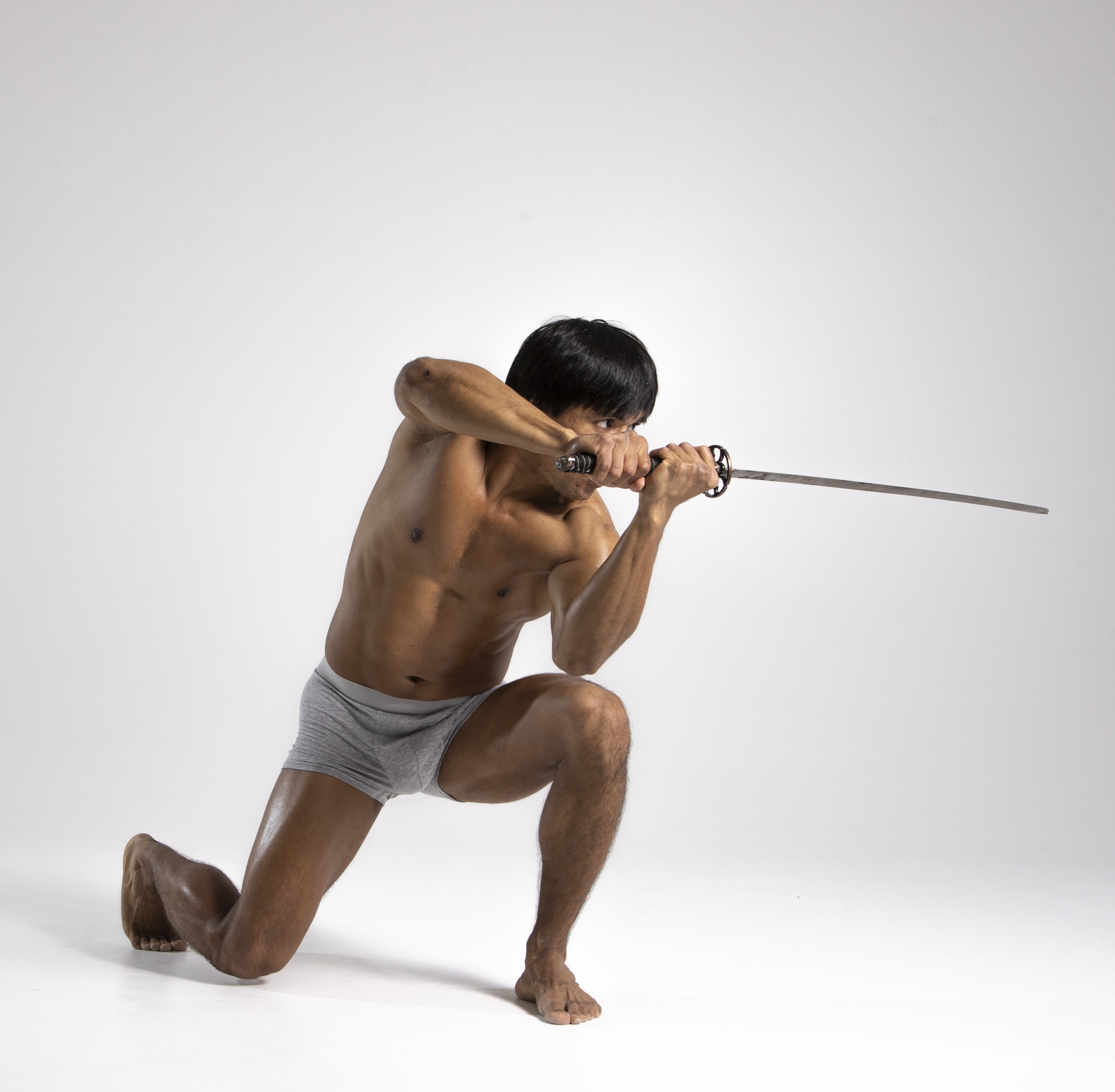 a shirtless asian man in gray underwear is kneeling barefoot and holding a katana sword in a studio shot against a white background. he is looking intently at the sword.