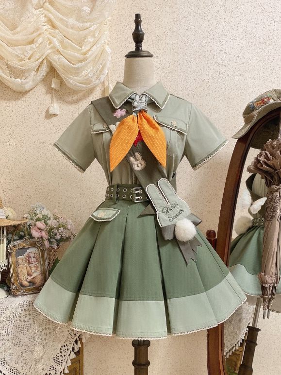 a green and brown lolita fashion outfit is displayed on a mannequin. the outfit consists of a collared shirt with embroidered bunnies, an orange necktie, a green skirt with brown accents and a belt, and a matching hat with bunny ears. the outfit is inspired by scouting and adventure.