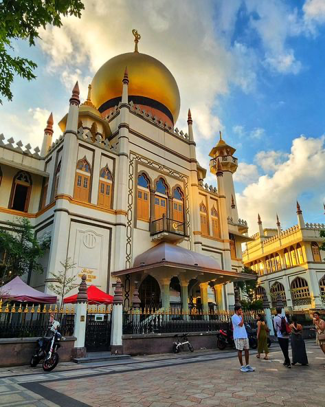 the image showcases the majestic masjid sultan in singapore, a prominent example of islamic architecture with a striking golden dome. the mosque's ornate facade, a blend of colonial and islamic styles, is prominently displayed. the photograph is taken from across the street, showing people walking around the mosque.  the bright afternoon sun casts shadows and highlights the building's architectural details. the scene is vibrant and captures a moment of daily life around this important religious site. the clear sky complements the building's grandeur.