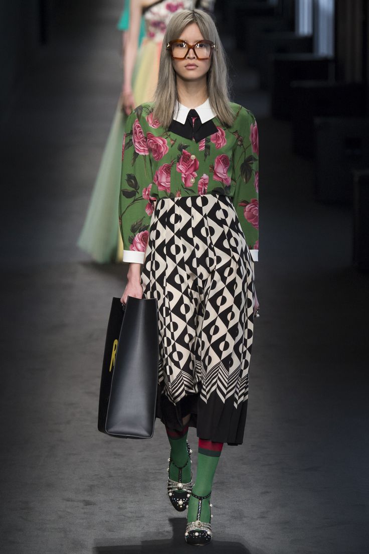 a runway model walks down the gucci runway wearing a green floral blouse paired with a black and white geometric midi skirt. she is accessorized with glasses, a black handbag, and jeweled shoes with green socks. the model has blonde hair and a composed expression. the background shows other models and a glimpse of the runway setting. the image is a highquality fashion photograph, likely from a professional fashion show.