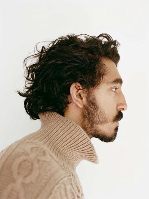 a profile shot of dev patel wearing a tan turtleneck sweater. he has curly dark hair, a beard, and a mustache. the background is white.