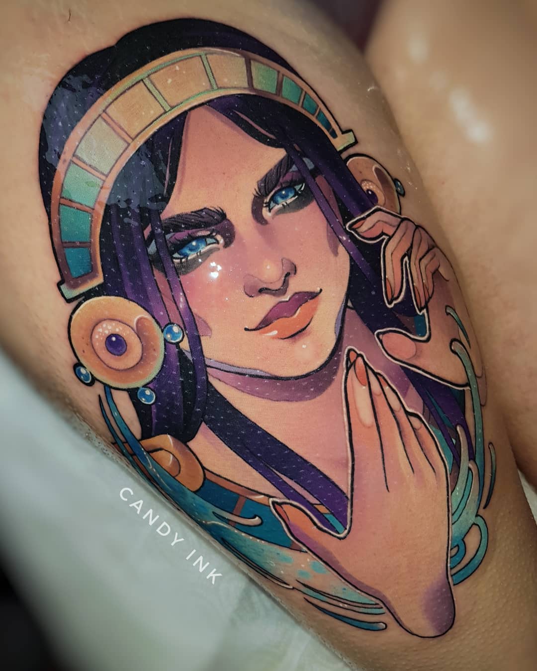a closeup image of a colorful tattoo of a woman on someone’s upper thigh. the woman in the tattoo has dark purple hair, blue eyes, and is wearing a large gold and teal headdress. she’s posed with one hand up by her face, and the other hand reaching out. the style of the tattoo is reminiscent of art nouveau.