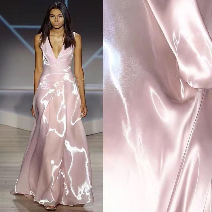 a model stands on a runway wearing a pink, floorlength gown. the gown has a vneck and is sleeveless. the fabric is sheer with a satin finish, creating a shiny and reflective surface. part of another piece of pink satin fabric is also in the image next to the model. the model is in the foreground and is the primary focus.