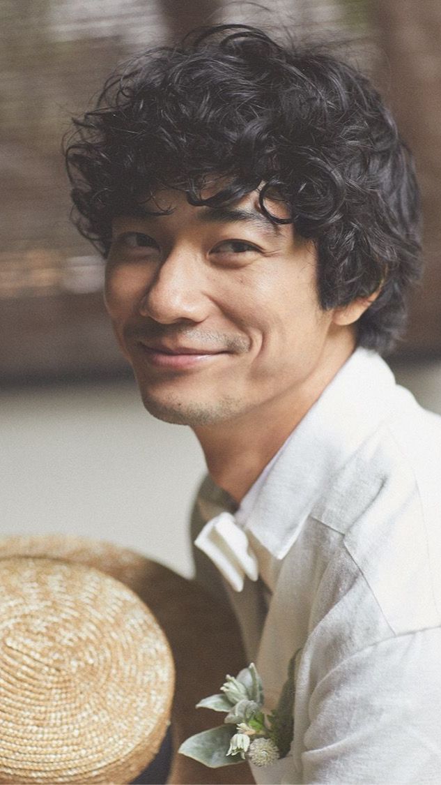 a closeup portrait of a smiling man with curly hair. he is wearing a white shirt and a straw hat. the man is looking at the camera with a relaxed and carefree expression. he has a flower pinned to his shirt. the image is likely taken outdoors.