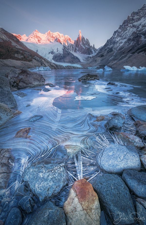 a landscape photograph features a partially frozen lake surrounded by rocks, with towering mountains in the background. the mountain peaks are touched by the warm glow of the rising sun, while the lake's surface displays intricate patterns of ice formations. the foreground is dominated by rocks and ice crystals that have formed around them. the overall scene captures the serene and rugged beauty of a winter landscape in patagonia, argentina, with the iconic cerro torre mountain visible in the distance.