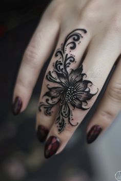 a hand with dark nail polish and a black flower tattoo on the middle finger. the tattoo is intricately designed with a flower as the central element and swirling patterns extending up the finger.