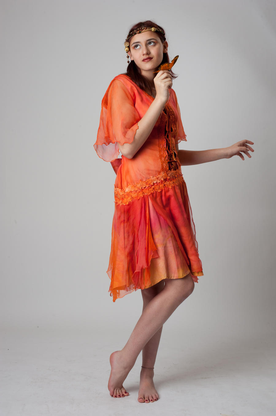 a fulllength shot of a young woman in an orange flowing dress. she is barefoot and holds a small object, possibly a decorative piece. the dress is sheer and has a layered look, with lace detailing near the waist. her expression is somewhat thoughtful, and the overall aesthetic is ethereal and artistic. the background is a simple light gray.