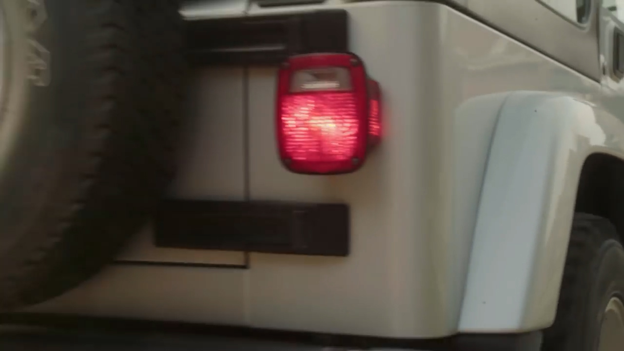 the image shows the rear view of a white jeep driving on a road. the red taillight of the jeep is illuminated. the environment appears to be daytime with natural, sunny lighting.