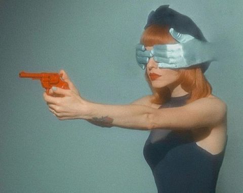 a fashionable woman with a blue beret and gloves over her eyes is holding a red toy gun.  she is pointing the gun towards the camera and has a serious expression on her face.  the  image is styled in a retro or vintage manner.