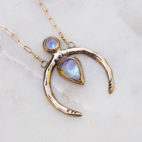 the image showcases a delicate gold necklace. the necklace features a crescent moon shaped pendant adorned with two moonstones. one is round, positioned at the top, while the other is teardropshaped, dangling from the center. the delicate gold chain of the necklace adds to its elegance, creating a harmonious blend of simplicity and ethereal beauty. the necklace is displayed on a white marble background, enhancing the piece's elegance. the soft, natural lighting used in the image highlights the iridescent shimmer of the moonstones and the intricate details of the gold pendant.