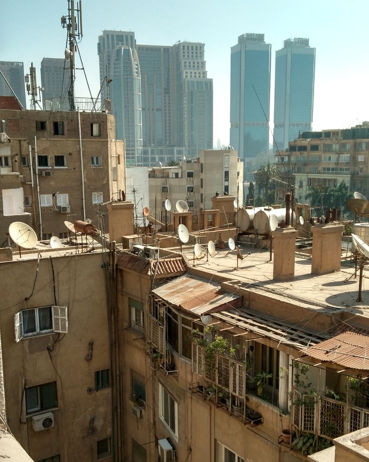 the image shows a view from a rooftop in cairo, egypt. in the foreground are older, multistory buildings with numerous satellite dishes and antennas on their rooftops. the buildings are primarily beige or brown, with balconies displaying some vegetation. the rooftops show signs of age and wear, including rusty metal. in the background, modern, glassfronted skyscrapers rise, creating a stark contrast between the older and newer architecture of the city. the overall impression is one of a dense and crowded urban environment.