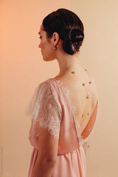 a woman in a pink, lacetrimmed dress stands with her back to the camera. her dark hair is styled in an elegant updo, and she wears a delicate gold necklace that drapes down her back. the warm, diffused studio lighting creates a soft and feminine atmosphere.