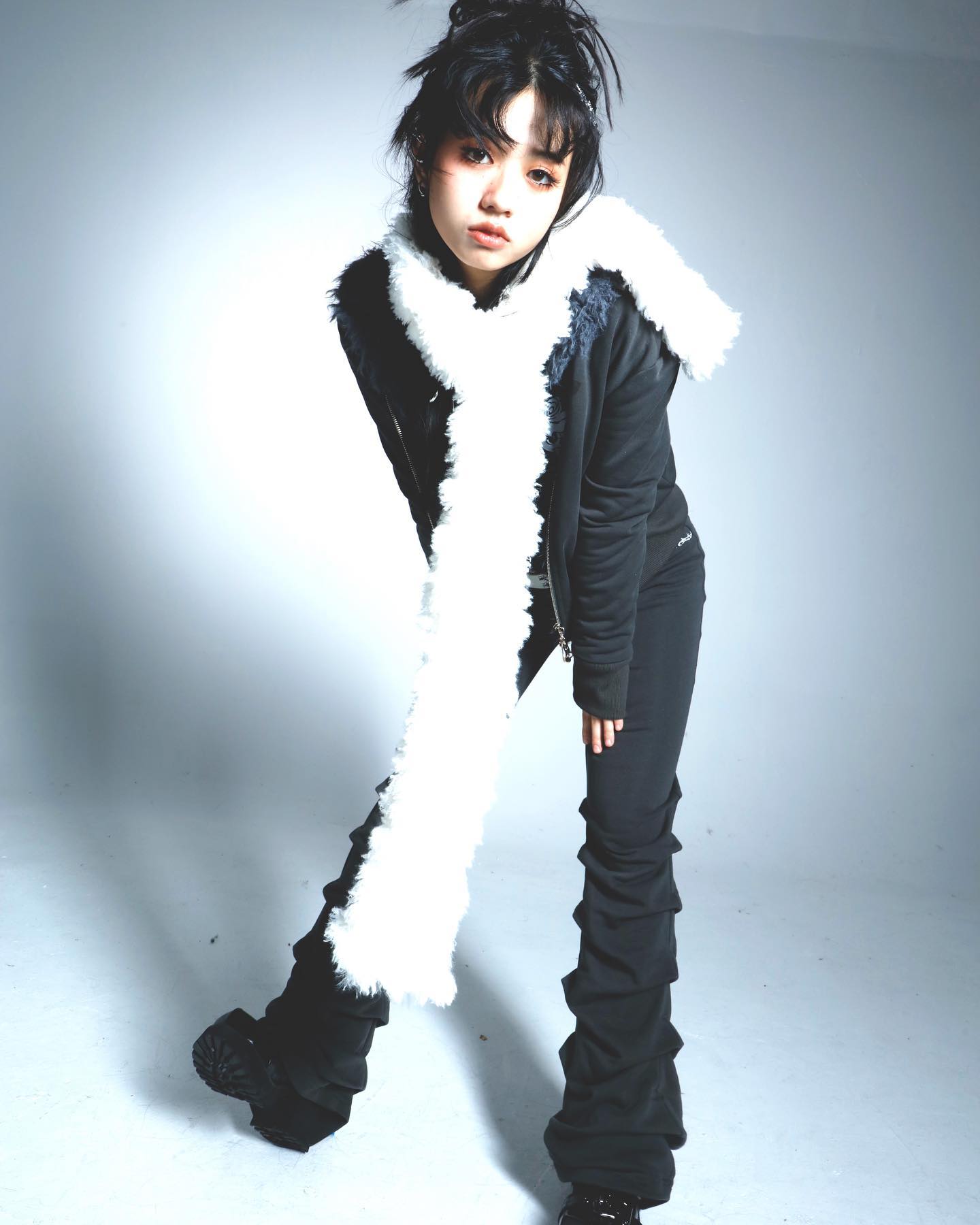 young asian woman with dark hair styled in a slightly messy updo is posing in a studio. she is wearing a black jacket, black pants with a gathered or pleated effect at the ankles, and a long white fluffy scarf.  her makeup is fairly neutral, and her overall style is dark and edgy, with a hint of grunge or alternative fashion.  the background is a plain light grey. the image is welllit and the focus is sharp.