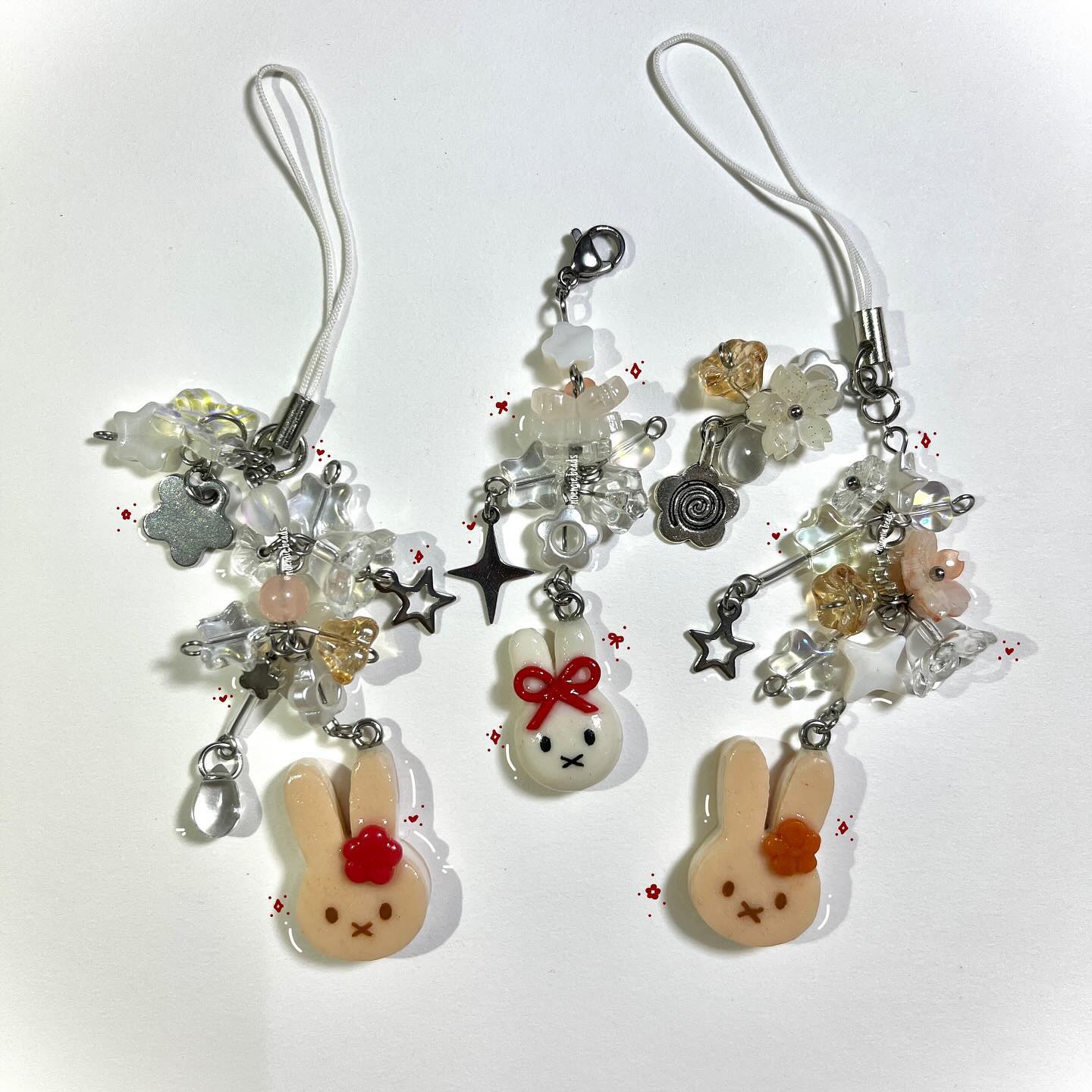 the image shows three handmade phone charms with a kawaii aesthetic. each charm features a bunny pendant at the bottom, various beads and metal charms (stars, flowers) strung together, and a white phone strap at the top. the charms have a delicate and handcrafted appearance, with a variety of bead colors and shapes.