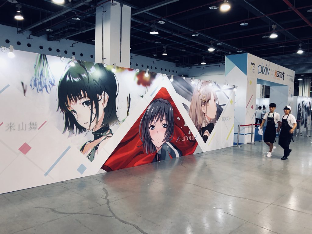 the image shows a large poster of anime characters at an anime convention. the poster is brightly lit and features three female characters. two people are walking by the poster. the overall color palette is white, black, and gray.  the floor is gray concrete and the ceiling is black.