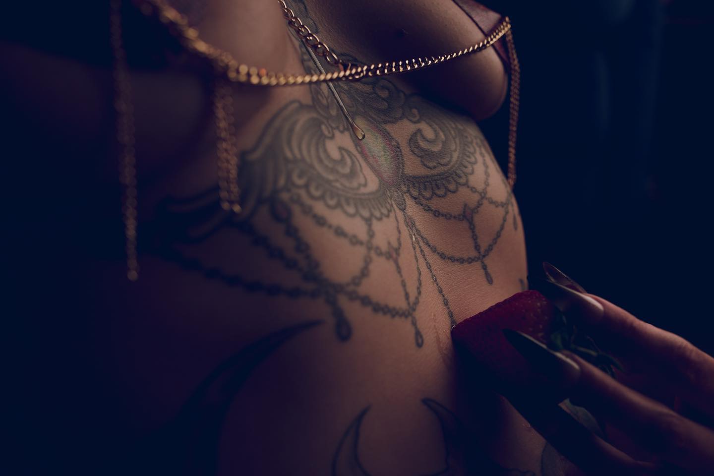 a closeup, sensual image of a woman's torso. the focus is on her intricately designed mandalastyle tattoo that spans across her chest and abdomen.  she is wearing a gold chainstyle lingerie and is holding a strawberry to her stomach with one hand. the lighting is low and moody, emphasizing the details of the tattoo and the woman's skin. the overall mood is intimate and suggestive.