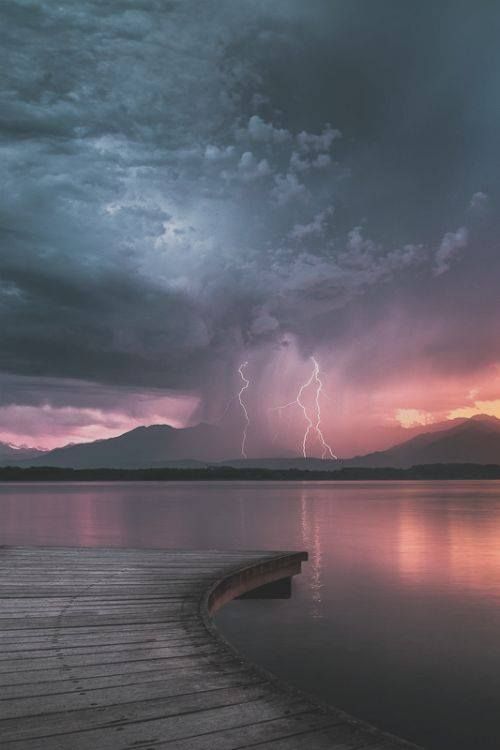 a wooden dock extends into a calm lake, surrounded by mountains. the sky is filled with dramatic storm clouds, illuminated by streaks of lightning. the sunset casts a warm glow over the scene, creating a moody and peaceful atmosphere.