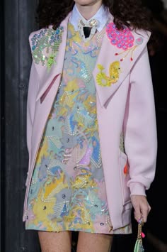 a woman model is wearing a pink jacket with an embroidered bodice and a colorful sequin dress. the look is very feminine and playful, with a touch of whimsy. she is posing for a photoshoot, and the lighting is bright and focused. the jacket is belted, and the model is wearing a statement necklace and earrings. she is wearing a stylish clutch bag that complements the overall look. the background is out of focus, allowing the model and her outfit to take center stage. the photo is wellcomposed, and the model is showcasing the outfit with confidence. the overall look is trendy and elegant, making it suitable for a party, event, or a special occasion.