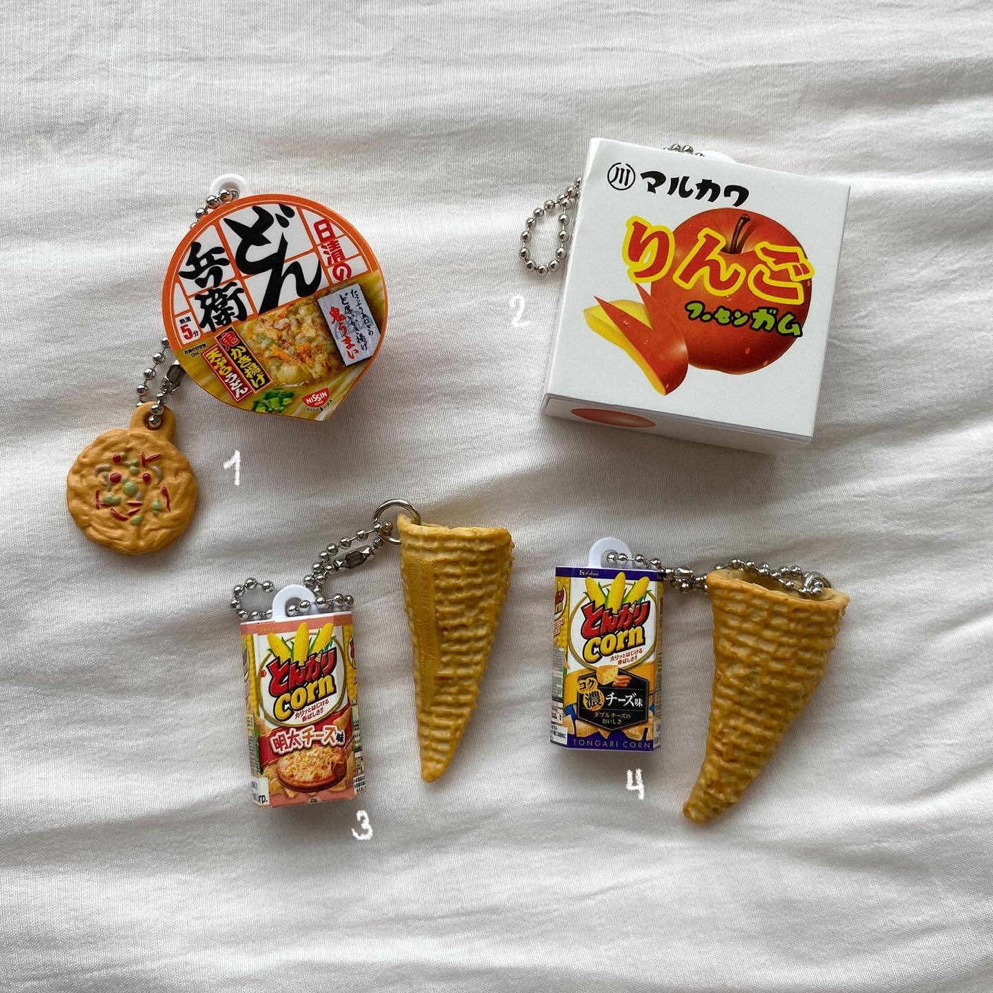 the image shows a collection of japanese snackthemed keychains arranged on a white, textured surface. the keychains feature miniature replicas of popular snacks, including nissin instant udon, marukawa apple gum, and tongari corn snack. each keychain is attached to a silver chain with a keyring. the items are labeled with numbers 14 to differentiate them.