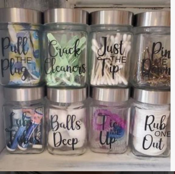 the image shows eight glass jars with silver lids, arranged in two rows of four, each labeled with a humorous phrase. the jars contain various bathroom or household items such as cotton swabs, bobby pins, razors, and cotton balls. the labels add a playful and slightly risque element to the storage containers.