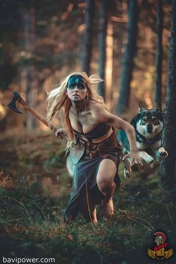 a woman dressed in a vikinginspired costume runs through a forest. she has war paint on her face, carries an axe, and is accompanied by a wolf running next to her.