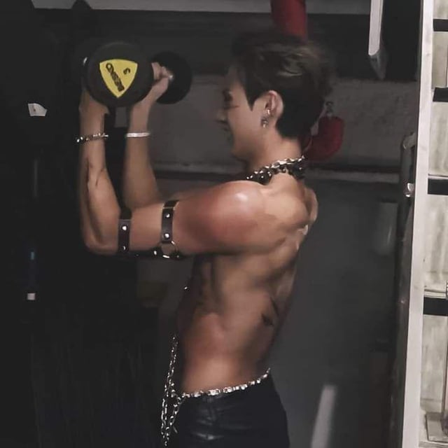 a shirtless muscular man is shown from the side, lifting a dumbbell. he is wearing dark leatherlike pants and various chains and accessories. the background is dark and dimly lit, possibly a gym or backstage area.  his physique is toned and welldefined, showcasing his muscular back and abs.  a small tattoo is visible on his lower back. the overall aesthetic is stylish and cool.