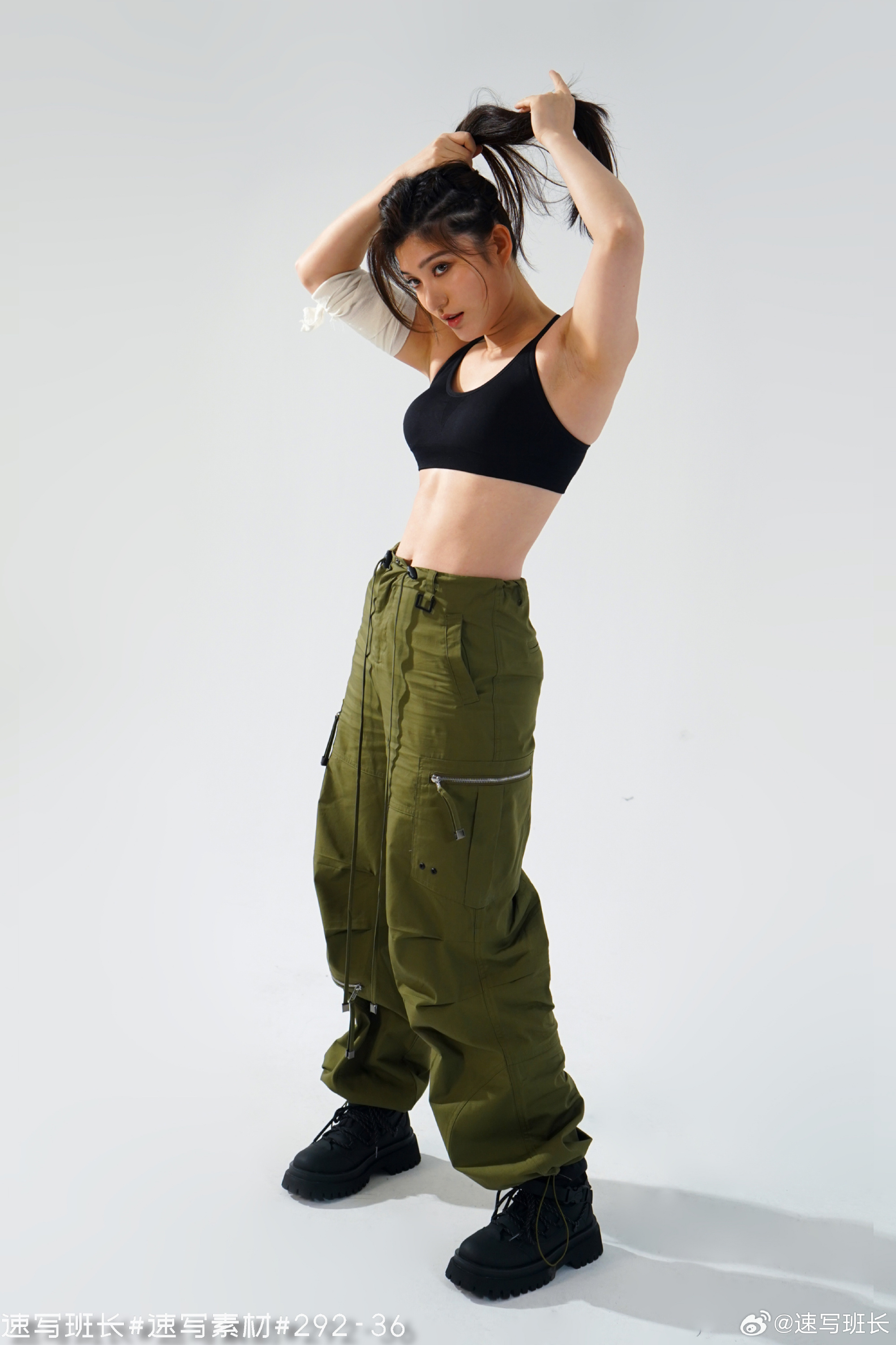 a woman stands posing in front of a white background, wearing a black sports bra and green cargo pants. she is tying her hair up in a ponytail. she has a white bandage wrapped around her left arm.