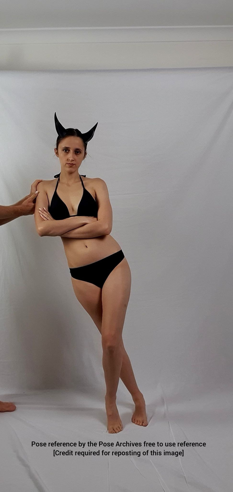 a woman in black swimwear and devil horns is standing in a contrapposto pose against a white background. her arms are crossed over her chest and she is looking directly at the viewer. a hand can be seen on her left arm, suggesting someone is guiding her pose.