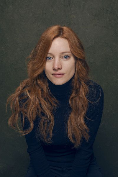 a woman with long, red hair and blue eyes is shown in a headshotstyle portrait. she is wearing a black turtleneck sweater and is posed against a dark green backdrop. her expression is neutral and her hair falls loosely around her shoulders. the lighting is soft and diffused, creating a natural and flattering look.