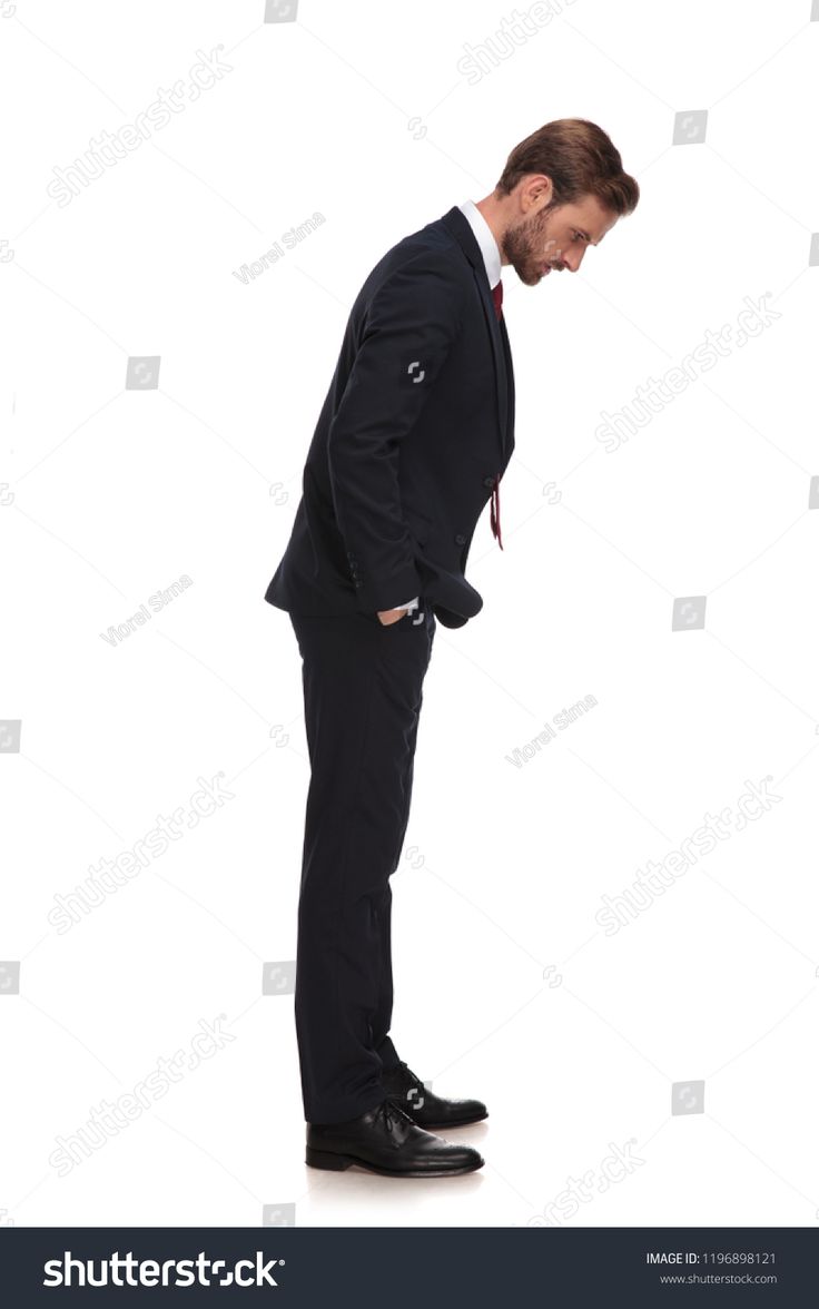 a man in a black suit is standing with his head down looking down at the ground. he is wearing a tie, a black suit jacket and black pants. he is wearing black shoes. he is standing on a white background.
