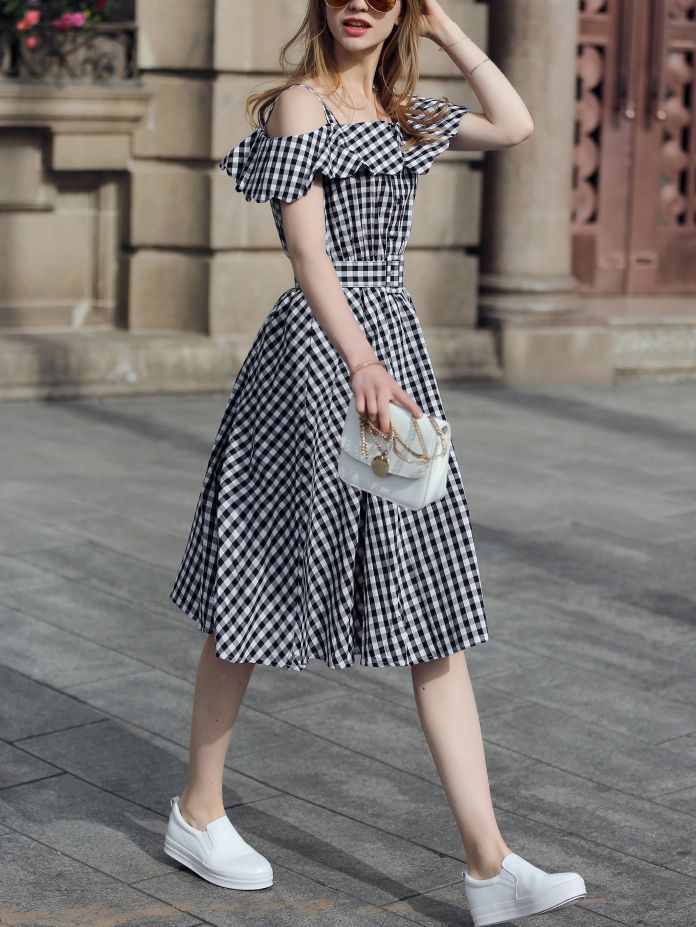 a young woman is wearing a black and white checkered offshoulder dress and white platform sneakers. she is carrying a white handbag with a gold chain strap and is wearing sunglasses. she is walking down a city street.