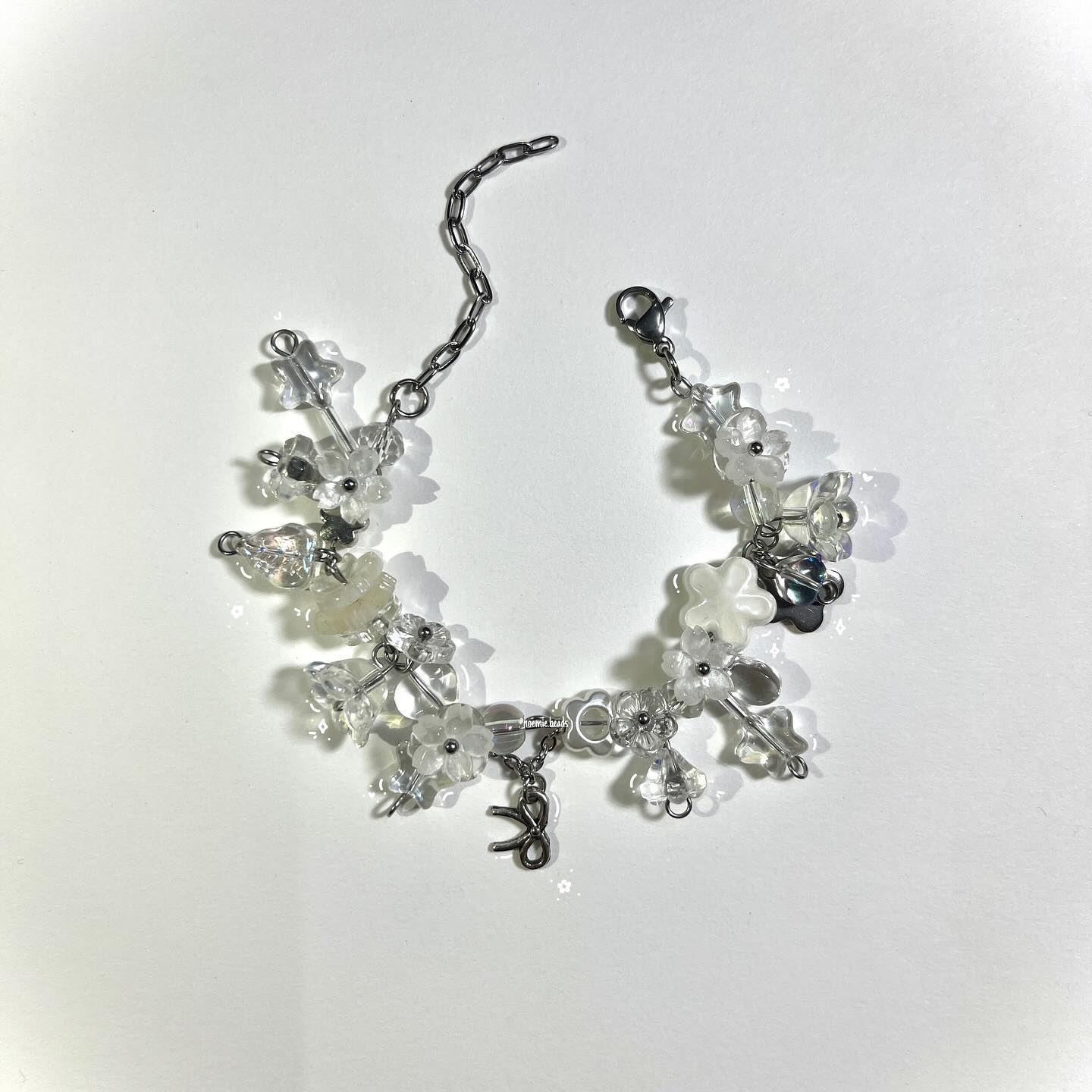 a bracelet with a silver chain adorned with clear beads and white floral beads. a small ribbon charm dangles from the bracelet. the bracelet is laying on a white background.