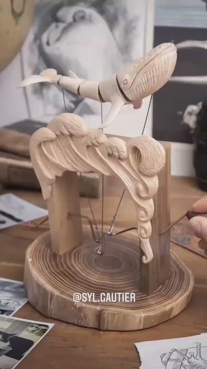 a wooden sculpture of a whale suspended above a carved wave. the sculpture is mounted on a round wooden base. the whale appears to be a marionette, connected by strings. the base has the watermark @syl.gautier.
