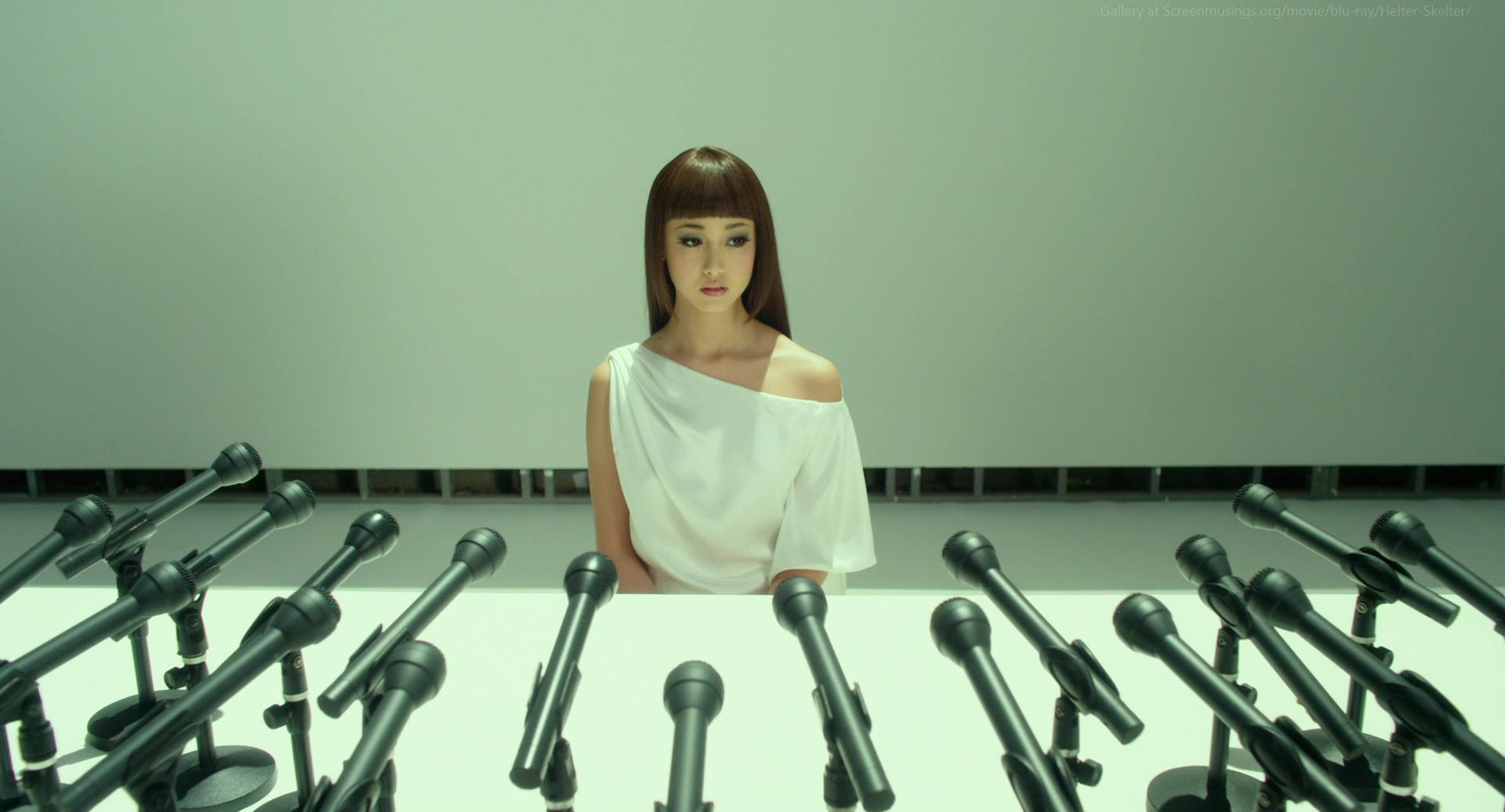a young woman in a white dress sits before a row of microphones, looking away from the camera with an apprehensive expression. the white background and the rows of microphones suggest she's at a press conference, perhaps waiting for a barrage of questions from reporters.  the overall mood is one of tension and anticipation.