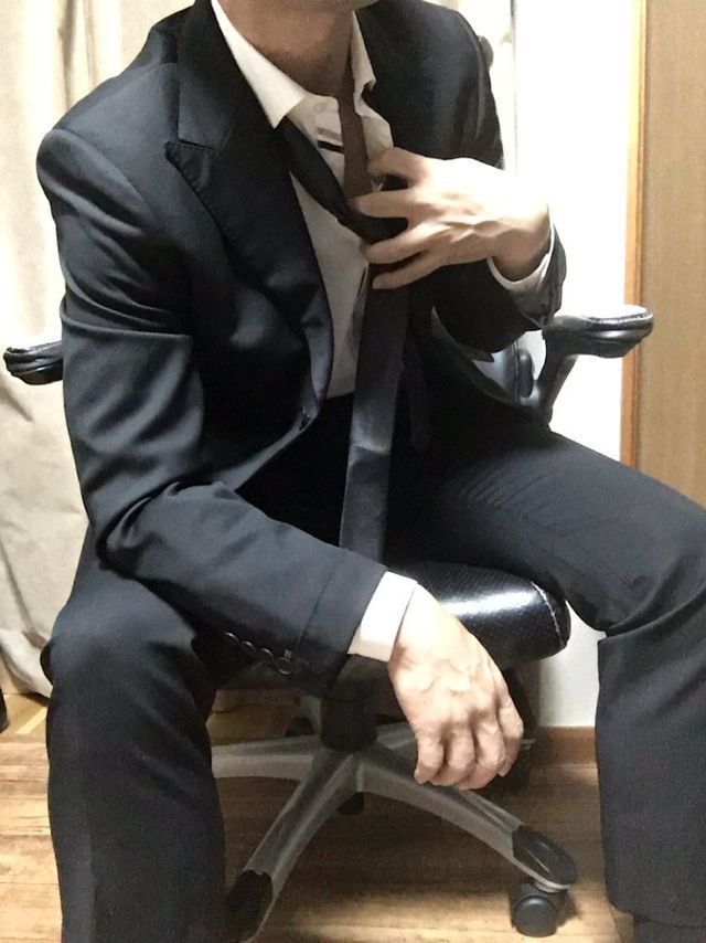 a man wearing a black suit and a white shirt is sitting on an office chair, with his hands resting on the armrest. the man is wearing a black tie, which is loose and hanging down his chest. the chair is black and has a silver base. the man is sitting in a relaxed manner. the background of the image is a wall, which is white.