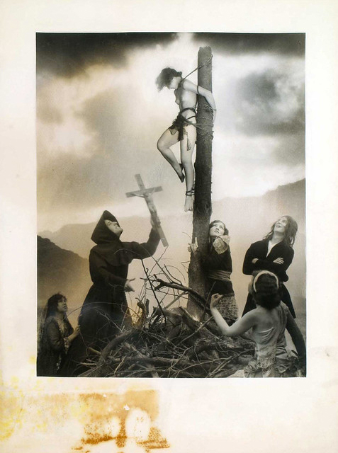 a black and white vintage photograph depicting a dramatic scene. a nude woman is tied to a wooden post, resembling a crucifixion. a hooded figure holds a cross aloft, seeming to preside over the event. several other women are present, some watching, some seemingly participating.  a bonfire is in the foreground. the setting appears to be outdoors in a mountainous area under a cloudy sky. the overall feel of the photo is dark, dramatic, and possibly surreal or symbolic. the style suggests it may be from the 1920s or early 20th century.  the photo is slightly damaged with a brown stain visible in the bottom left corner.