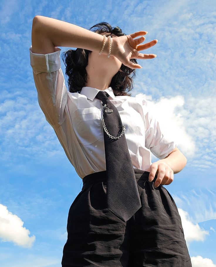 a lowangle, full shot of a young woman wearing a white shirt and black shorts, standing outdoors under a bright blue sky with fluffy white clouds. she is shielding her eyes from the sun with her hand and looking up. she is wearing a black tie with a chain and pendant accessory. the overall style is fashionable and slightly androgynous. the image evokes a sense of carefree happiness and youthful energy.