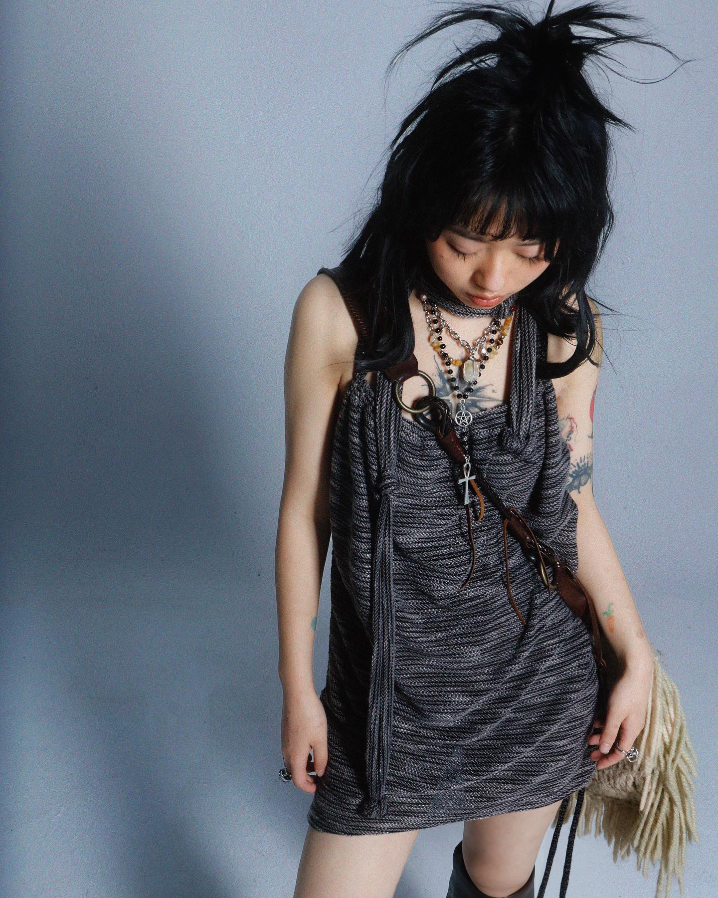 a young woman with dark hair styled in a messy bun is shown in a studio shot. she is wearing a dark grey knitted sleeveless dress with long shoulder straps and several necklaces, including a cross necklace. she also has bracelets and rings on. she is looking down, and a part of her tattoo is visible on her arm. the background is a solid light grey.