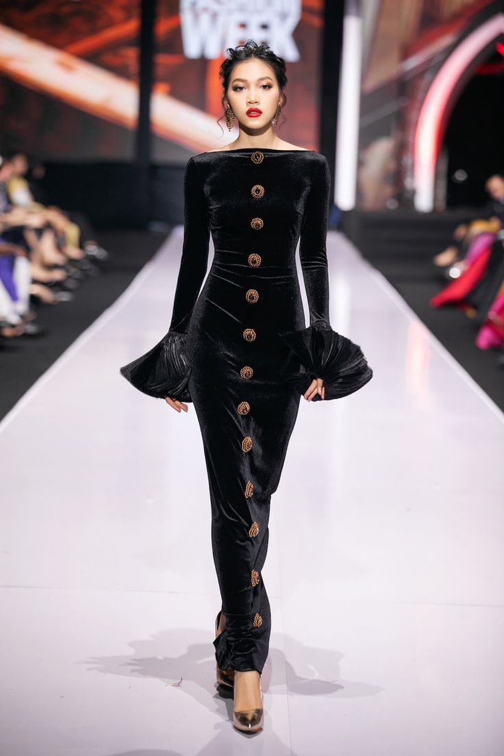 a full shot of a model walking down a runway in a black velvet dress. the dress has long sleeves with bellshaped cuffs and a row of decorative buttons down the front. the model is wearing high heels and has her hair pulled back. the background is a bright white runway with a blurred background suggesting a fashion show.