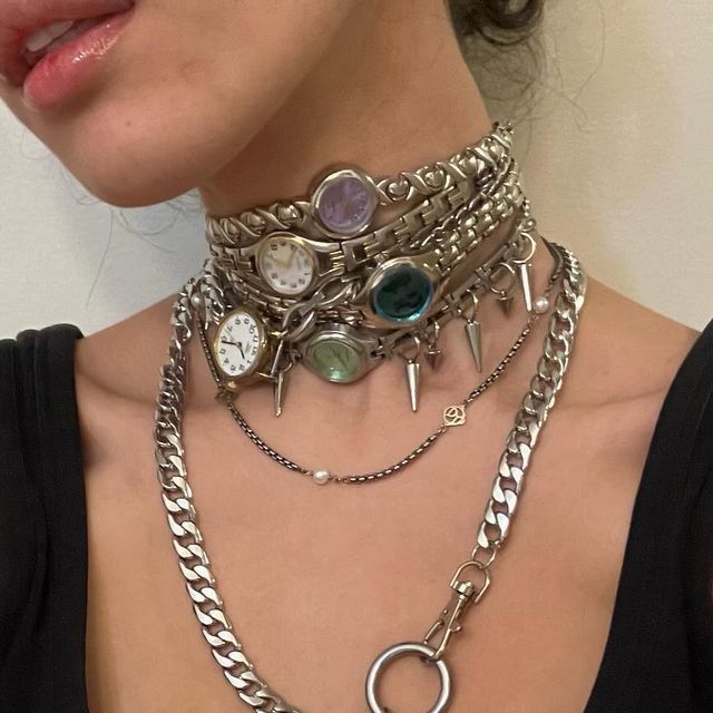 loseup shot of a person wearing a layered necklace consisting of several silver chains and multiple small watches attached as decorative elements. the chokerstyle necklace is adorned with metal spikes and some gemstones, giving it a punk or grunge aesthetic.  a longer, heavy silver chain necklace is layered over the watch choker, with a delicate chain and a small pendant visible underneath. the overall style is edgy and fashionable.