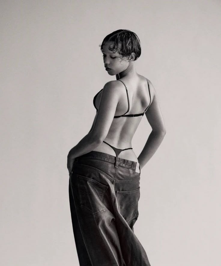 a woman is wearing a bra and pants, and she is looking down with her hands in her pockets. it is a black and white portrait.