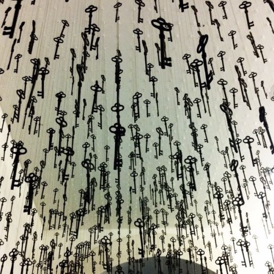 the image shows a large number of black key silhouettes hanging from a white ceiling. the keys are various sizes and arranged in a pattern, creating an art installation. the perspective is looking up, giving the impression of depth and a multitude of hanging objects. the shadows of the keys add to the visual effect.